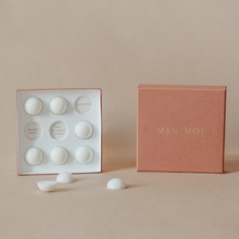 An open masmoi box revealing floral scent massage oil pearls inside, each designed to melt into nourishing oil for a soothing massage. The inner packaging includes playful text such as 'I got your back' and 'head goosebumps incoming,' adding a personalised touch. Set against a warm beige background, this luxurious product is perfect for a massage workshop or self-care ritual.